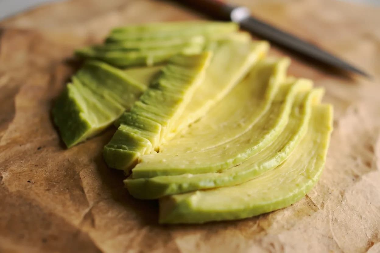 Avocado Fan Cut in its raw whole-cut form on a butcher's board — The avocado fan cut involves halving the avocado lengthwise, removing the pit, peeling or scooping the flesh, making a series of parallel lengthwise cuts through the widest part while leaving the nar… Identifying traits: Intact hinge at stem end keeps all slices connected as a single plating unit, Fan geometry displays color gradient from center to edge on a flat plate, Parallel cuts create uniform slice thickness (4–6 mm) for even eating.